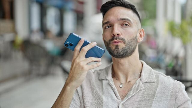 Young hispanic man sending voice message by smartphone sitting on table at coffee shop terrace