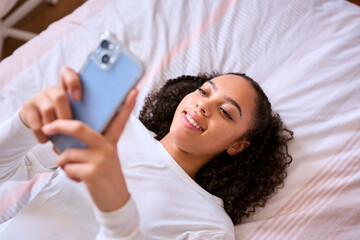 Teenage Girl At Home Lying On Bed Connecting With Friends On Social Media Using Mobile Phone