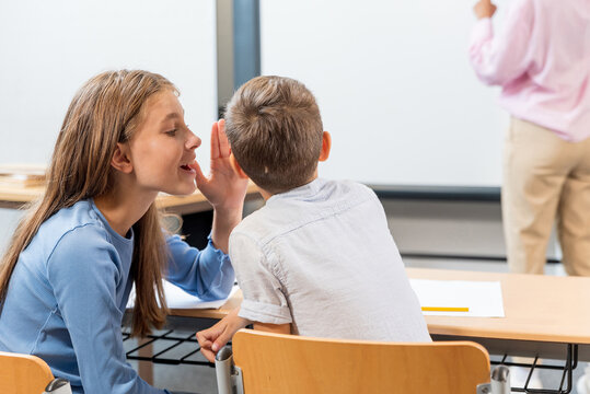 Schoolchildren, friends and whispering in classroom for secret, gossip or chatting during teaching lesson. learners sharing quiet information secretly in ear at class while teacher is busy. - Powered by Adobe