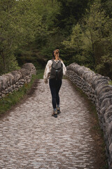 Young woman with backpack hiking and crossing an old stone bridge in the middle of the forest