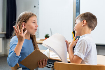 Lively conversation of children in the classroom. A schoolgirl explains to her desk mate how letters are written. The boy is interested and thoughtful. Creative communication of children as teacher.