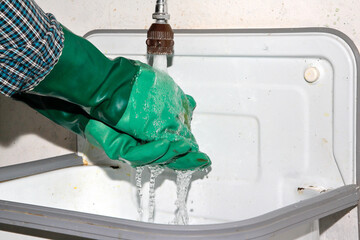 A man puts his hands with green rubber gloves under running water. Beware of greenwashing! True sustainability requires more than just cleaning colored gloves.