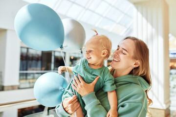 Portrait of happy mother with baby in her arms and colored balloons. Young pretty mom and her little daughter in same green clothes walk around the mall and have fun. Family weekend in shopping mall.
