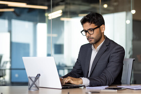 Young serious and focused businessman at workplace typing on computer, Arab man in business suit sitting at table thinking about solving technical work tasks.