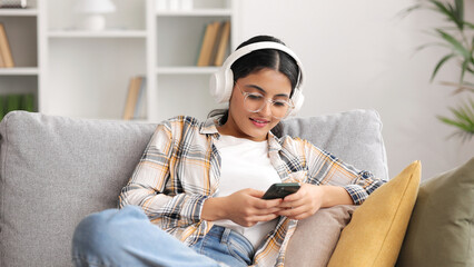 Happy young girl with white headphones chilling and listening her favorite song. Indian brunette woman seat on sofa and relax after work, uses smartphone for play music, clicking on the screen.