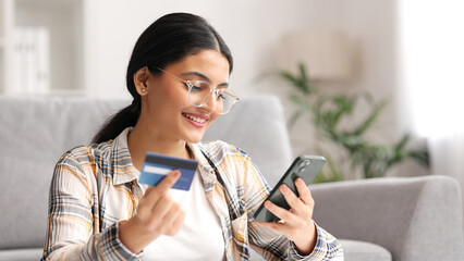 Smiling pretty young Indian brunette girl holding mobile phone and credit card sitting at the table. Happy woman makes online shopping using mobile payment. E-banking app service.