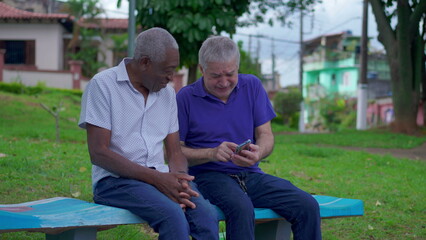 Two diverse seniors looking at phone sitting on park bench outdoors laughing and smiling together. Older friend sharing online media content to companion depicting authentic friendship