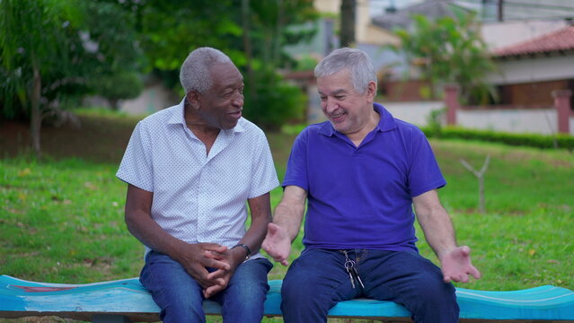 Two diverse senior talking and laughing sitting on park bench in Brazil. Candid authentic people real life laugh and smile in old age retirement golden years