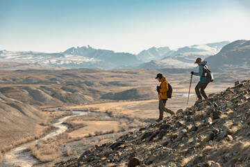 Two young hikers with backpacks and hiking poles walk downhill in mountains. Active tourism in...