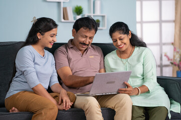 Happy indian middle aged parents with adult daughter using laptop on sofa at home - concept of Modern Family, Togetherness and Parental Support