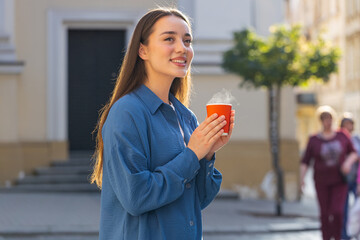 Fototapeta premium Portrait of pretty young woman enjoying morning aroma coffee steam or tea hot drink outdoors. Relaxing, taking a break. Lovely girl walking in urban city sunny street sun rays, drinking coffee to go