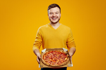 Excited man in yellow sweater holding cardboard flat box with pizza isolated on orange background. Food delivery concept