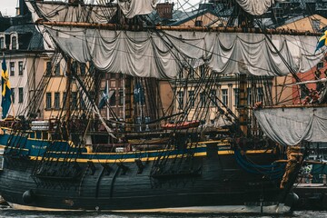 large wooden ship in the water near a city, with buildings and sky background