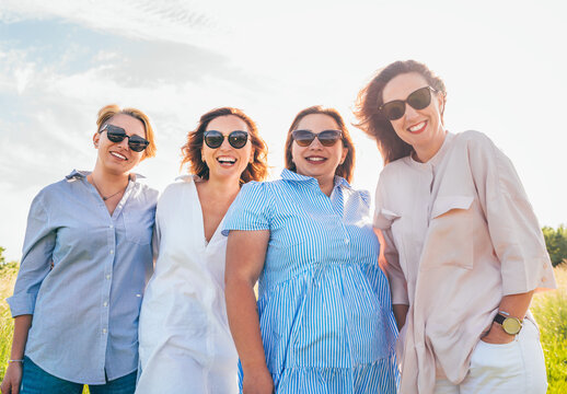 Portrait of four cheerful smiling women in sunglasses embracing and looking at the camera during outdoor walking. Woman's friendship, relations, and happiness concept image.