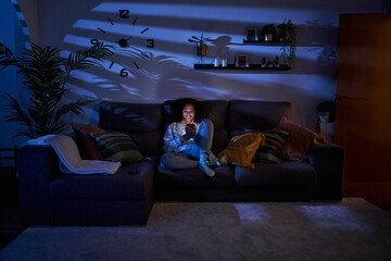 Young woman with happy expression looking at cell phone sitting in living room at night. Smiling cheerful latina female using mobile on sofa at home. Copy space, generation z and free time indoors.