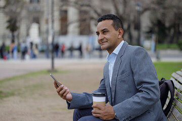 Hispanic man with a disposable cup of coffee sitting on a bench in a public park.