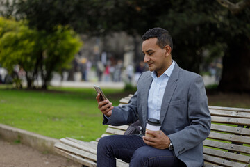 Fototapeta premium Hispanic man in a suit and a disposable cup of coffee sitting on a bench in a public park.