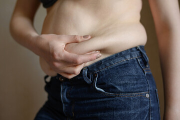 Naklejka premium A fat woman in jeans with a protruding belly touches the fat folds on her stomach, close-up .Weight loss, obesity.