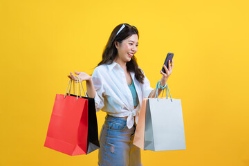 happy shopper with smartphone smiling and holding shopping bags isolated on yellow studio...
