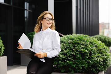Elegant confident young woman, formally dressed, sits outdoors against the background of the business center, holding a folder with documents, looks at clock - obrazy, fototapety, plakaty