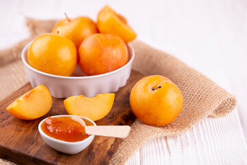 Close-up of plum jam in a white bowl and yellow plums whole and pieces on a wooden board on burlap on the table. Copy space.