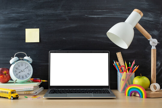 Education theme. Side view photo of desk with stationery, notebooks, laptop, alarm clock, plasticine, apples, lamp and school-related props on chalkboard backdrop. Perfect for text or ad placement