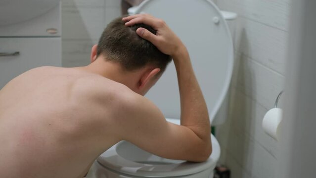 A man sits leaning towards the toilet feeling nauseous, rear view