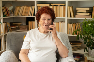 Portrait of a modern elderly woman who sits in an armchair against the background of a bookcase with books and a flower, pleasantly talking on the phone, smiling and looking at the camera.