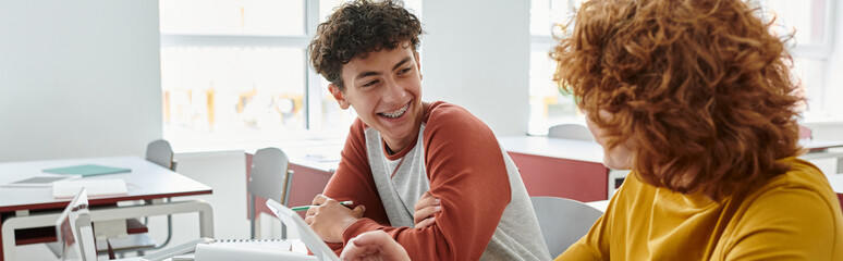 Smiling teen schoolboy talking to classmate near devices during lesson in classroom, banner