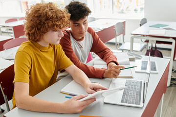 Teenage schoolboys using devices together during lesson in classroom at background in school