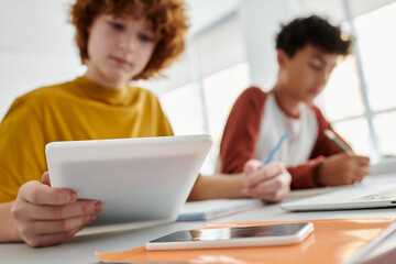 Blurred teen schoolboy using digital tablet near friend and notebook during lesson in classroom