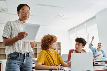 Obraz premium African american teacher holding digital tablet during lesson near schoolboys in classroom