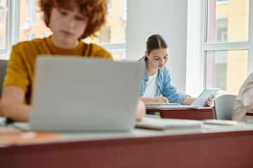 Fototapeta na wymiar Teen schoolgirl holding digital tablet and writing on notebook near blurred classmate in classroom
