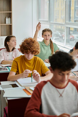 Obraz premium Teenage schoolgirl raising hand and talking near devices and classmates during lesson in school