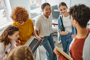 redhead boy showing laptop to classmate, african american teacher smiling near students