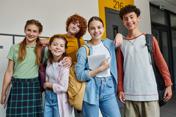 back to school, happy teenagers looking at camera in hallway, holding devices and smiling together