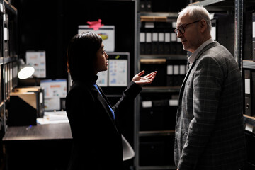 Accountancy asian assistant reporting budget plan optimizations to elderly businessman executive manager. Dark file cabinet office room surrounded by document folders on cabinet shelves
