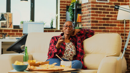 Happy female adult laughing at comedy film on tv, relaxing on couch with beer and snacks. Young...