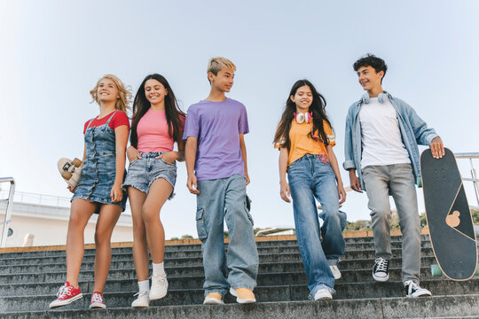 Group of smiling friends wearing colorful t shirts talking, communication, walking on the street with skate boards. Happy stylish boys and girls outdoors. Friendship, positive lifestyle, summer