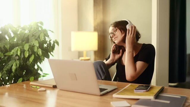 Energetic young woman freelancer with headphones listening music while working online typing browsing on laptop and dancing indoors Positive female enjoying great working day at cozy home