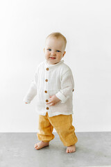 One year old caucasian blonde boy dressed in white and nude urban clothes looking at camera in a white background