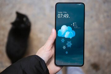 Woman checking weather using app on smartphone outdoors, closeup. Data and illustration of cloud with snowflakes on screen