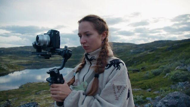 Caucasian young girl with brown hair in a light jacket with pigtails turns on the stabilizer for shooting a summer landscape with a lake and hills on a stabilizer and camera on a cloudy day in Norway