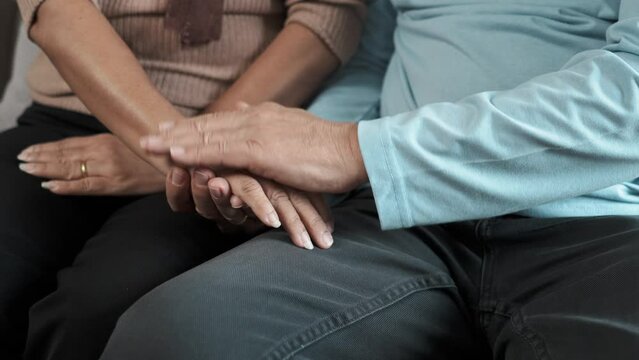 Close up view of senior couple holding hands while sitting on the sofa in the living room together.