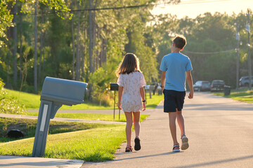Back view of two young teenage children, girl and boy, brother and sister walking together on suburban street on bright sunny evening