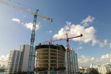 Fototapeta premium Tower lifting cranes at high residential apartment building construction site. Real estate development in Miami urban area