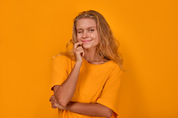 Young cute coquettish Caucasian woman student with long hair winks eye for flirts with you and looking at screen or trying to seduce dressed in oversize t-shirt stands on isolated yellow background.