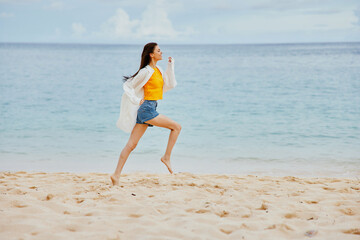 Fototapeta premium Sports woman runs along the beach in summer clothes on the sand in a yellow T-shirt and denim shorts white shirt flying hair ocean view