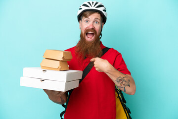 Delivery man holding pizzas and burgers isolated on blue background with surprise facial expression