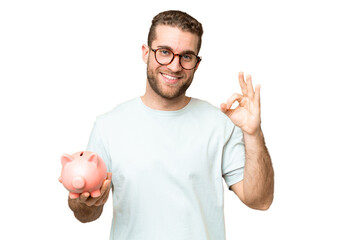 Young man holding a piggybank over isolated chroma key background showing ok sign with fingers